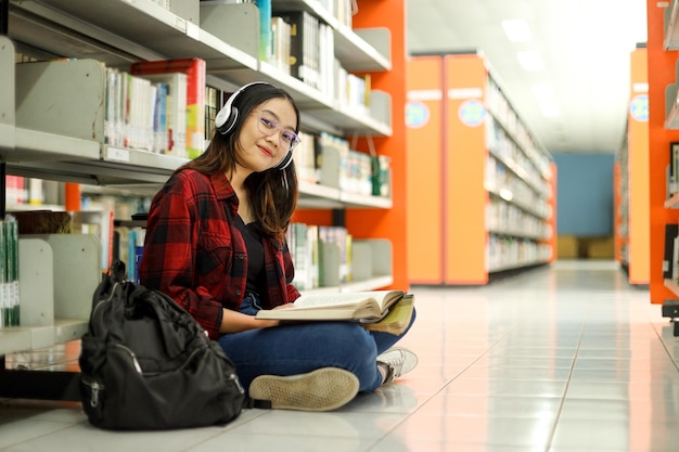 Alegre joven estudiante asiática mirando la cámara sonriendo y sentada con las piernas cruzadas en la biblioteca mientras