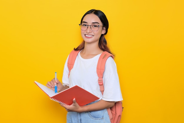 Alegre joven estudiante con mochila y anteojos escribiendo en un cuaderno aislado de fondo amarillo