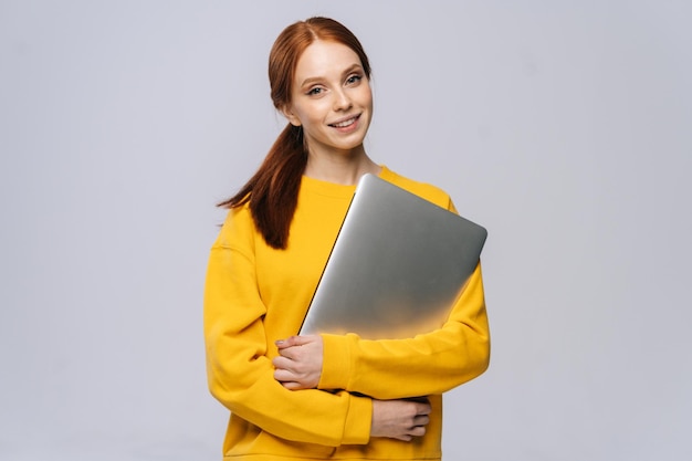 Alegre joven estudiante sosteniendo una computadora portátil cerrada y mirando a la cámara sobre un fondo blanco aislado Bonita modelo de dama con el pelo rojo mostrando emocionalmente expresiones faciales en el estudio