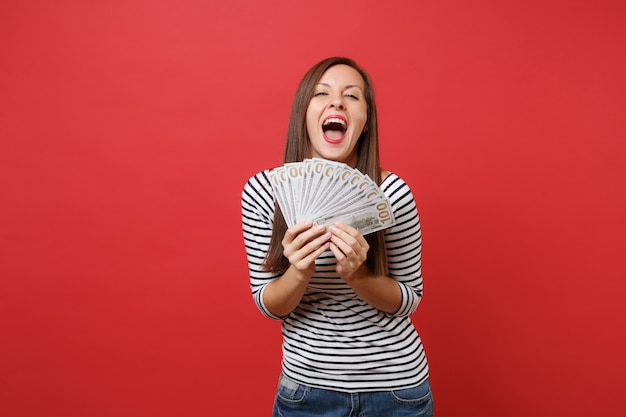 Alegre joven feliz manteniendo la boca abierta, sosteniendo un montón de dólares, dinero en efectivo aislado sobre fondo rojo brillante de la pared. Personas sinceras emociones, concepto de estilo de vida. Simulacros de espacio de copia.