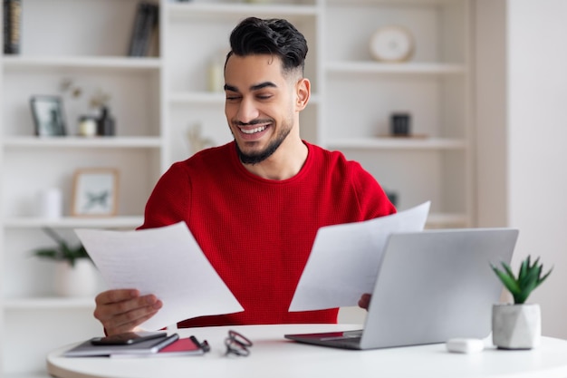 Alegre joven gerente del medio oriente con barba trabaja con documentos en el lugar de trabajo con computadora