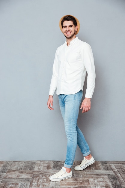 Alegre joven guapo con camisa blanca, jeans y sombrero sonriendo y caminando