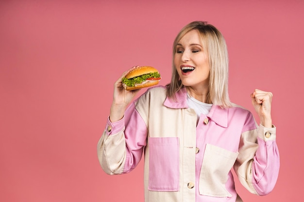 Alegre joven hambrienta hermosa mujer rubia sosteniendo una hamburguesa con queso o una hamburguesa en las manos aisladas sobre fondo rosa. Concepto de comida chatarra.