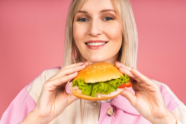 Alegre joven hambrienta hermosa mujer rubia sosteniendo una hamburguesa con queso o una hamburguesa en las manos aisladas sobre fondo rosa. Concepto de comida chatarra.