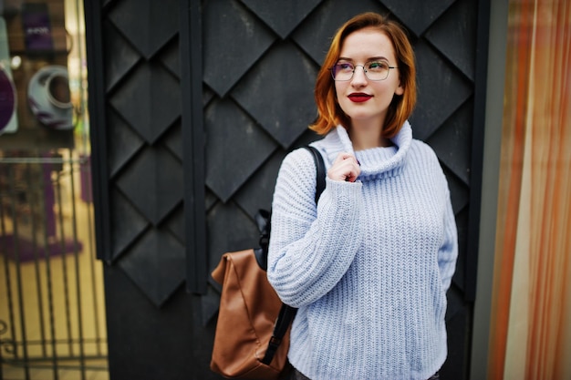 Alegre joven hermosa mujer pelirroja en gafas cálido suéter de lana azul con mochila posada al aire libre