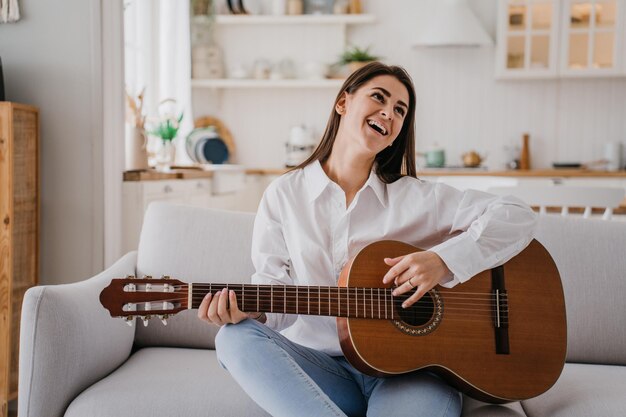 Foto alegre joven hispana tocando la guitarra en casa vestida con camisa blanca y jeans bastante