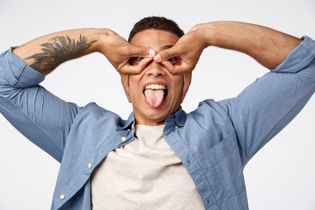 Alegre joven hispano guapo en camisa azul sobre camiseta, mostrando la lengua felizmente