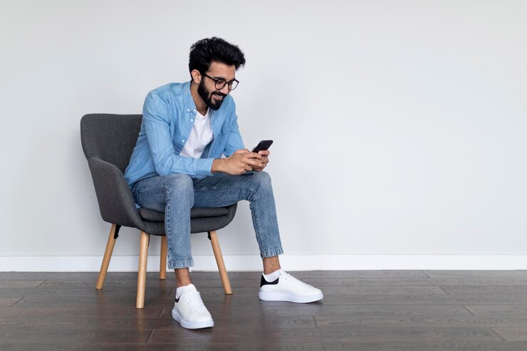 Alegre joven indio usando teléfono celular mientras está sentado en un sillón en casa