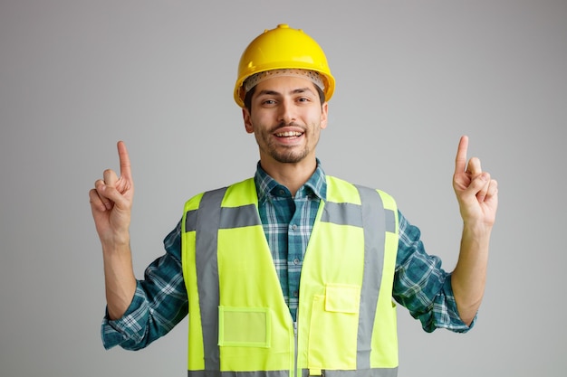 Alegre joven ingeniero masculino con casco de seguridad y uniforme mirando a la cámara apuntando hacia arriba aislado en fondo blanco