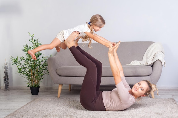 Alegre joven madre practicando yoga con su pequeña hija criando a un niño divirtiéndose y mirando a la cámara familia feliz haciendo ejercicio de gimnasia juntos entrenamiento físico con el niño en la salud del hogar