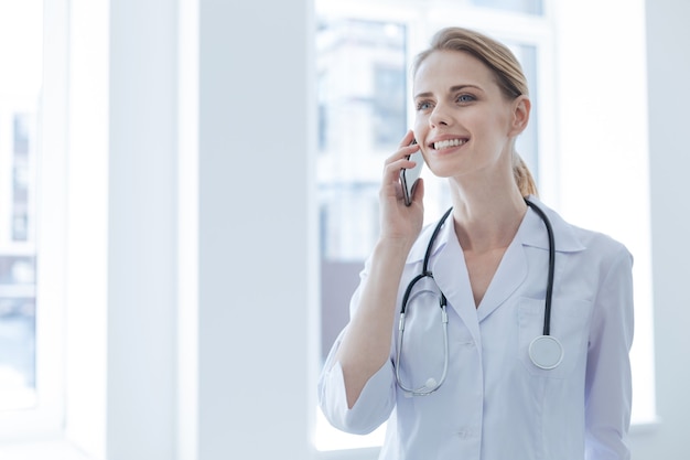 Alegre joven médico encantado disfrutando de las horas de trabajo en el hospital mientras expresa alegría y conversa por teléfono