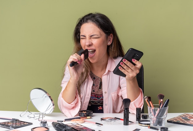 Alegre joven morena sentada a la mesa con herramientas de maquillaje sosteniendo teléfono y peine fingiendo cantar