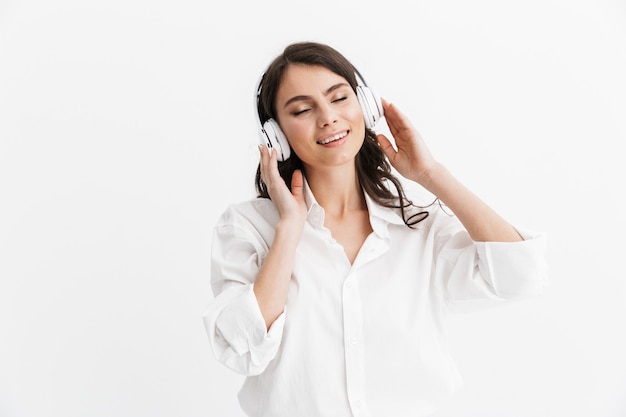 Alegre joven morena vistiendo camisa que se encuentran aisladas sobre la pared blanca, escuchando música con auriculares