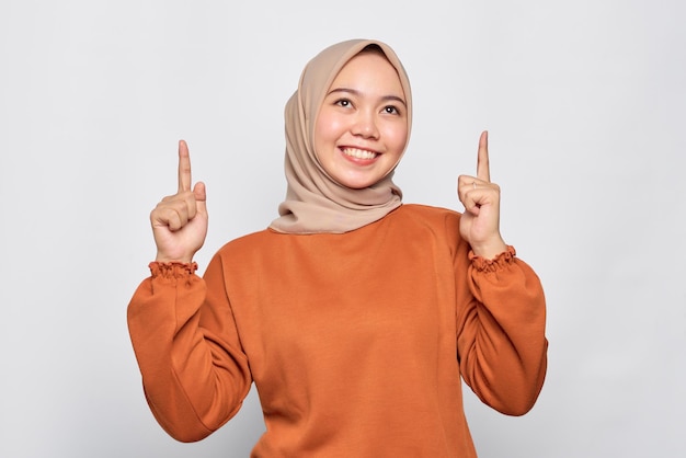 Alegre joven mujer asiática en camisa naranja apuntando con el dedo hacia arriba en el espacio de copia aislado sobre fondo blanco.