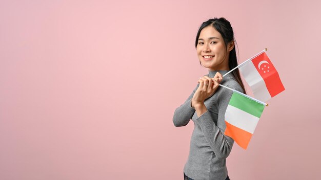 Alegre joven mujer asiática mostrando una bandera de Singapur e Irlanda sobre fondo rosa aislado