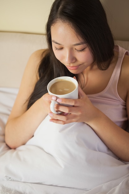 Alegre joven mujer asiática sentada en la cama que huele su café de la mañana