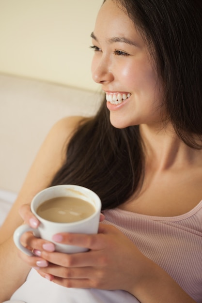 Alegre joven mujer asiática sentada en la cama con su café de la mañana
