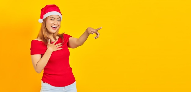 Alegre joven mujer asiática con sombrero de navidad apuntando con el dedo a la cámara sobre fondo amarillo