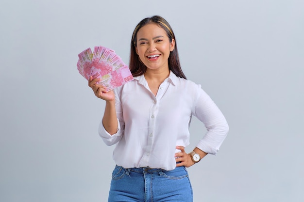 Alegre joven mujer asiática sosteniendo billetes de dinero y mirando a la cámara aislada sobre fondo blanco.