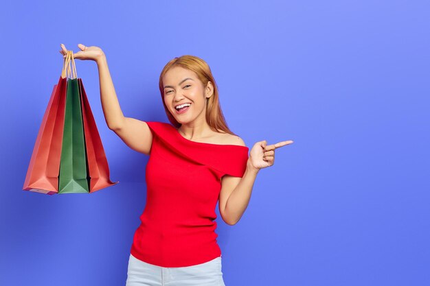 Alegre joven mujer asiática sosteniendo bolsas de compras y señalando con el dedo el espacio de copia aislado sobre fondo púrpura