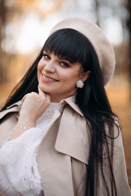 Alegre joven mujer caucásica con cabello largo oscuro en blusa blanca, pantalón gris, sombrero blanco y abrigo gris con bolso marrón en el bosque