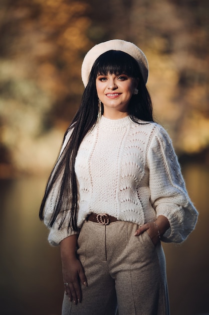 Alegre joven mujer caucásica con cabello largo oscuro en blusa blanca, pantalón gris, sombrero blanco y abrigo gris con bolso marrón camina cerca del río