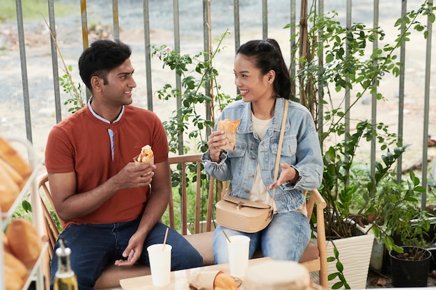 Alegre joven y mujer comiendo sándwiches y hablando al aire libre