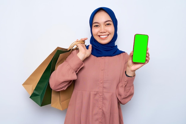 Alegre joven mujer musulmana asiática en camisa rosa sosteniendo bolsas de paquetes con compras después de ir de compras y mostrando el teléfono móvil de pantalla en blanco aislado sobre fondo blanco Concepto de estilo de vida de la gente