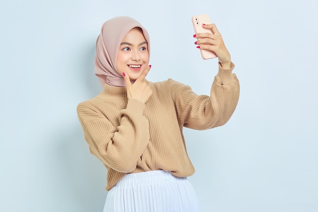 Alegre joven mujer musulmana asiática en suéter marrón haciendo selfie tiro en teléfono móvil aislado sobre fondo blanco Concepto de estilo de vida religioso de la gente
