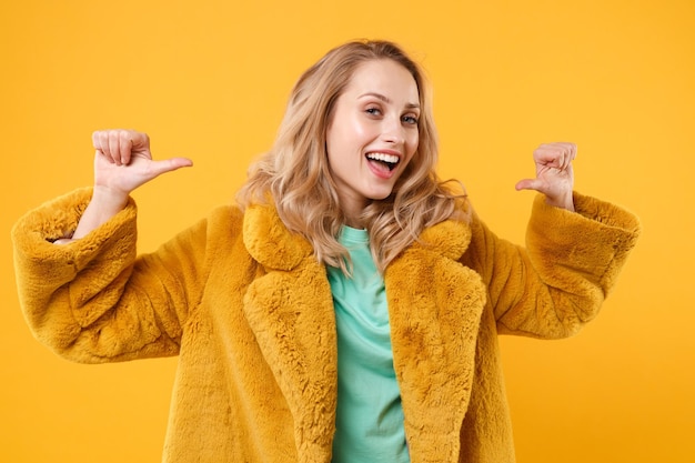 Alegre joven mujer rubia con abrigo de piel amarillo posando aislada en un retrato de estudio de fondo naranja. Gente emociones sinceras concepto de estilo de vida. Simulacros de espacio de copia. Apuntando los pulgares sobre sí misma.