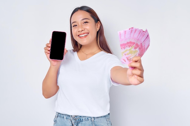 Alegre joven mujer rubia asiática con camiseta blanca casual que muestra el teléfono móvil con pantalla en blanco y con billetes de dinero en rupias aislados en el concepto de estilo de vida de la gente de fondo blanco