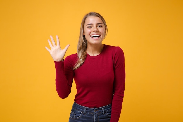 Alegre joven mujer rubia con ropa informal posando aislada en un fondo amarillo anaranjado en el estudio. Concepto de estilo de vida de las personas. Simulacros de espacio de copia. Agitando el saludo con la mano cuando alguien se da cuenta.