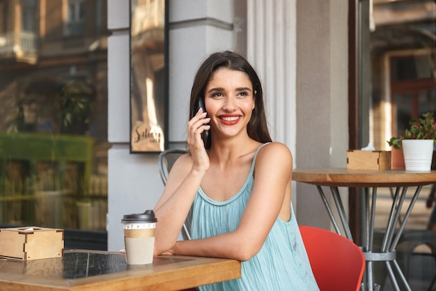 Alegre joven mujer sentada en la cafetería al aire libre mientras habla por teléfono móvil tomando café