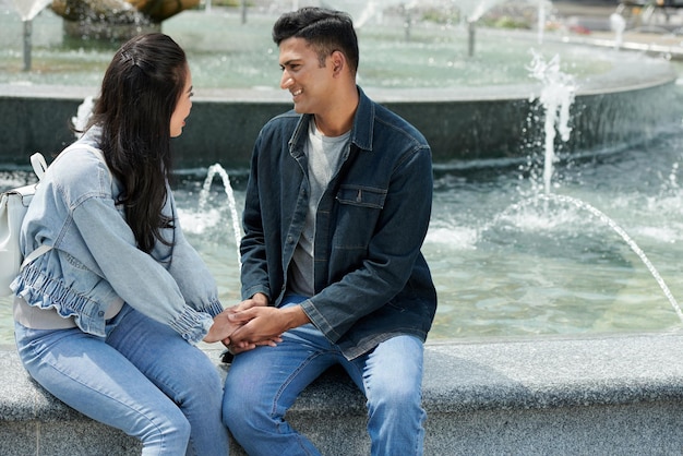 Alegre joven y mujer sentados en la fuente cogidos de la mano hablando y mirándose