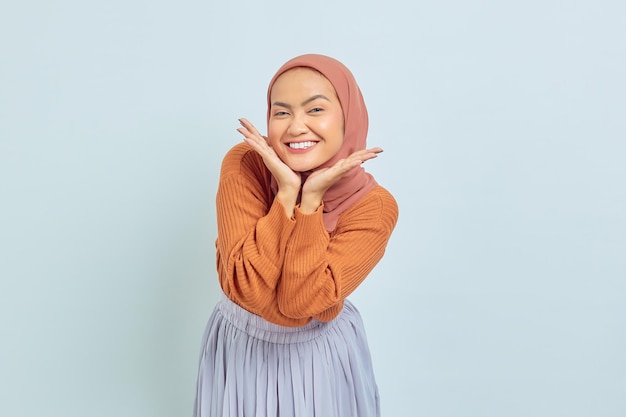 Alegre joven musulmana asiática con suéter marrón y poniendo las palmas en la barbilla aislada sobre el concepto de estilo de vida musulmán de fondo blanco