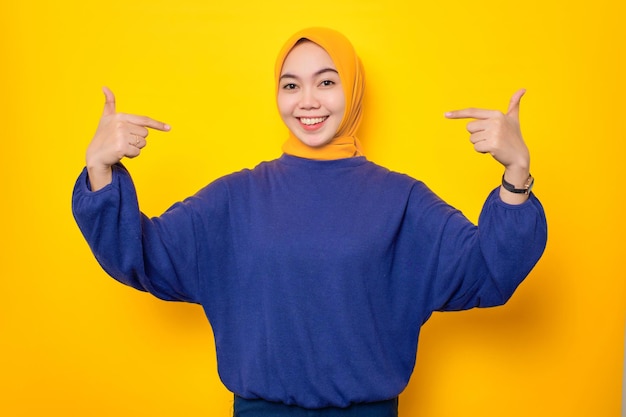 Una alegre joven musulmana asiática vestida con un suéter informal apuntándose a sí misma con orgullo aislada de un fondo amarillo