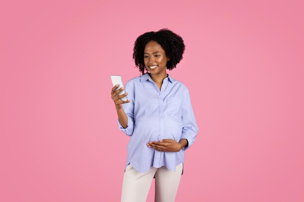 Alegre joven negra embarazada con un toque casual en el vientre usa un teléfono inteligente charlando en las redes sociales