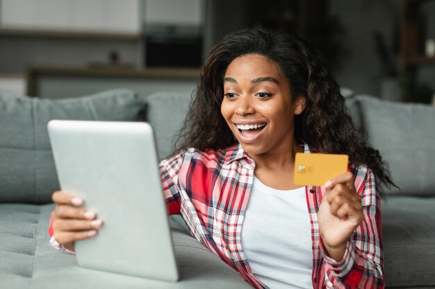 Alegre joven negra sorprendida mostrando tarjeta de crédito y tableta para compras en línea en la sala de estar