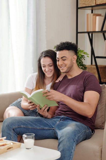 Alegre joven novio y novia sentados en el sofá con el libro en las manos