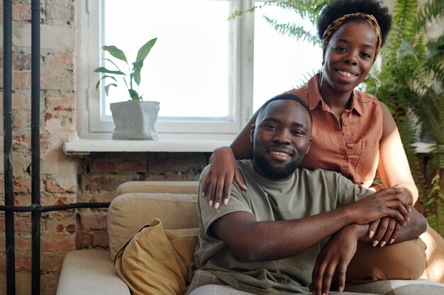 Alegre joven pareja africana en ropa casual mirándote con una sonrisa mientras está sentado frente a la cámara contra la ventana con plantas domésticas