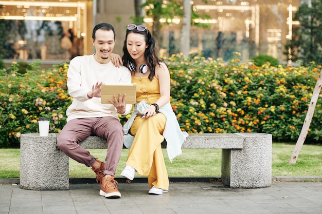 Alegre joven pareja china sentada en un banco y discutiendo videos o fotos en la computadora de la tableta
