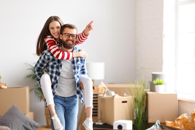 Alegre joven pareja familiar dando un paseo a cuestas a una mujer emocionada mientras se divierten juntos en la habitación con cajas con cosas después de la reubicación en un nuevo apartamento