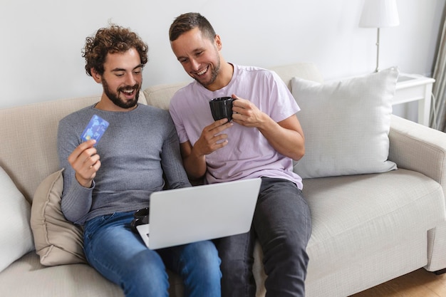 Alegre joven pareja gay sonriendo alegremente mientras compra en línea en casa Dos jóvenes amantes masculinos usando una tarjeta de crédito y una computadora portátil para hacer una compra en línea Joven pareja gay sentados juntos en el interior