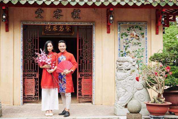 Alegre joven pareja vietnamita en trajes tradicionales de pie en las puertas del templo con ramas de melocotón en flor y abanico de papel