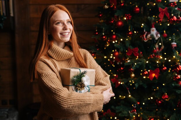 Alegre joven pelirroja feliz sosteniendo hermosas cajas de regalo de Navidad en el fondo del árbol de Navidad