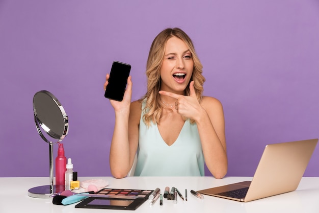 alegre joven positiva con ordenador portátil y cosméticos con espejo apuntando mostrando la pantalla del teléfono móvil.