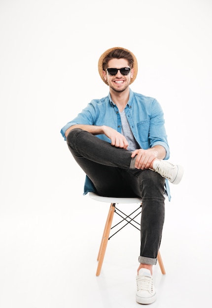 Alegre joven relajado con sombrero y gafas sentado en una silla sobre fondo blanco.