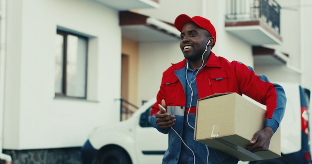 Alegre joven repartidor afroamericano en uniforme rojo sacando la caja de cartón de una camioneta y caminando hacia la casa mientras escucha la música en los auriculares y sonriendo. Al aire libre.