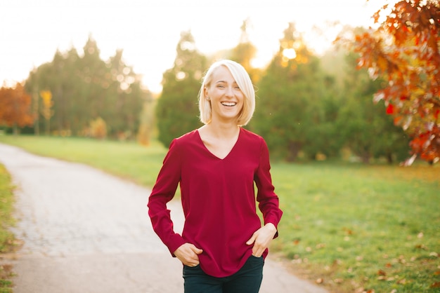 Alegre joven rubia, caminando en el parque otoño, sonriendo y metiendo las manos en los bolsillos