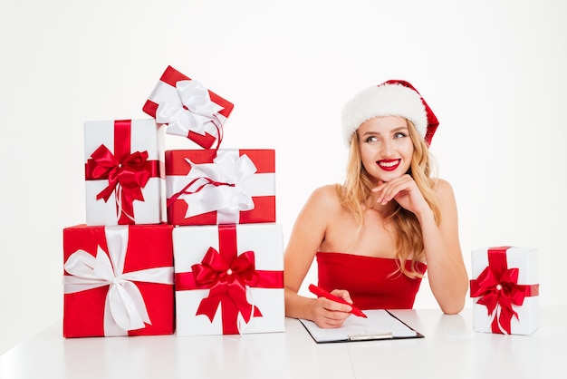 Alegre joven rubia escribiendo en un portapapeles con una gran cantidad de cajas de regalo